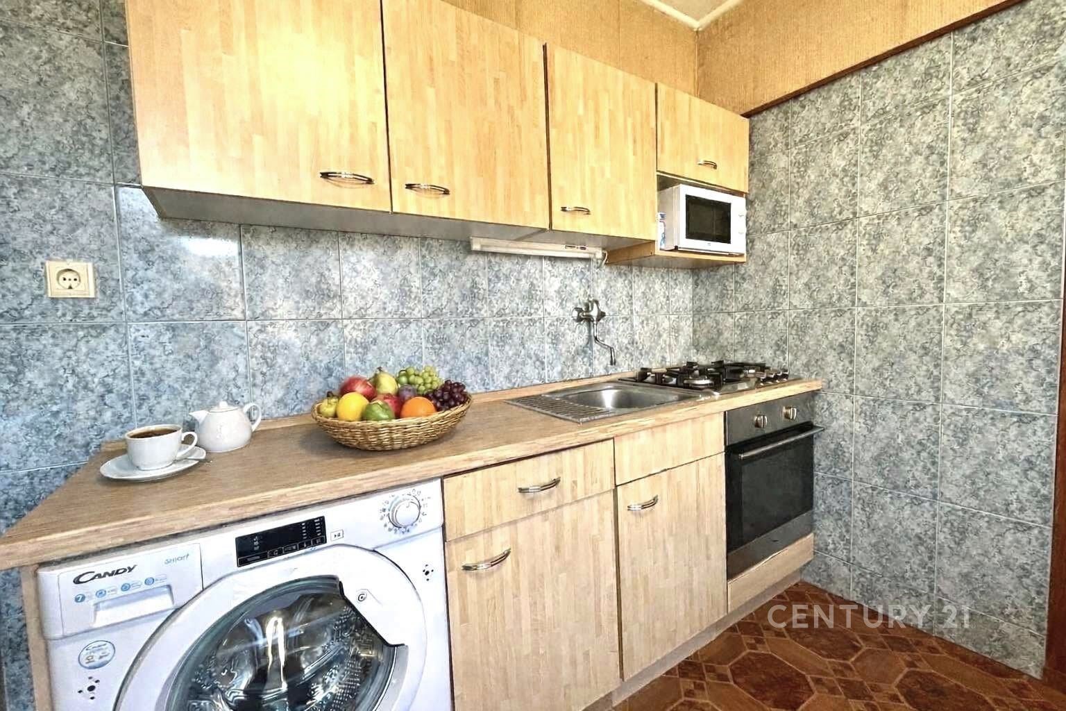 Kompaktní kuchyň s dřevěnými skříňkami, dlažbou a vestavěnými spotřebiči. Obsahuje pračku a malou mikrovlnnou troubu.