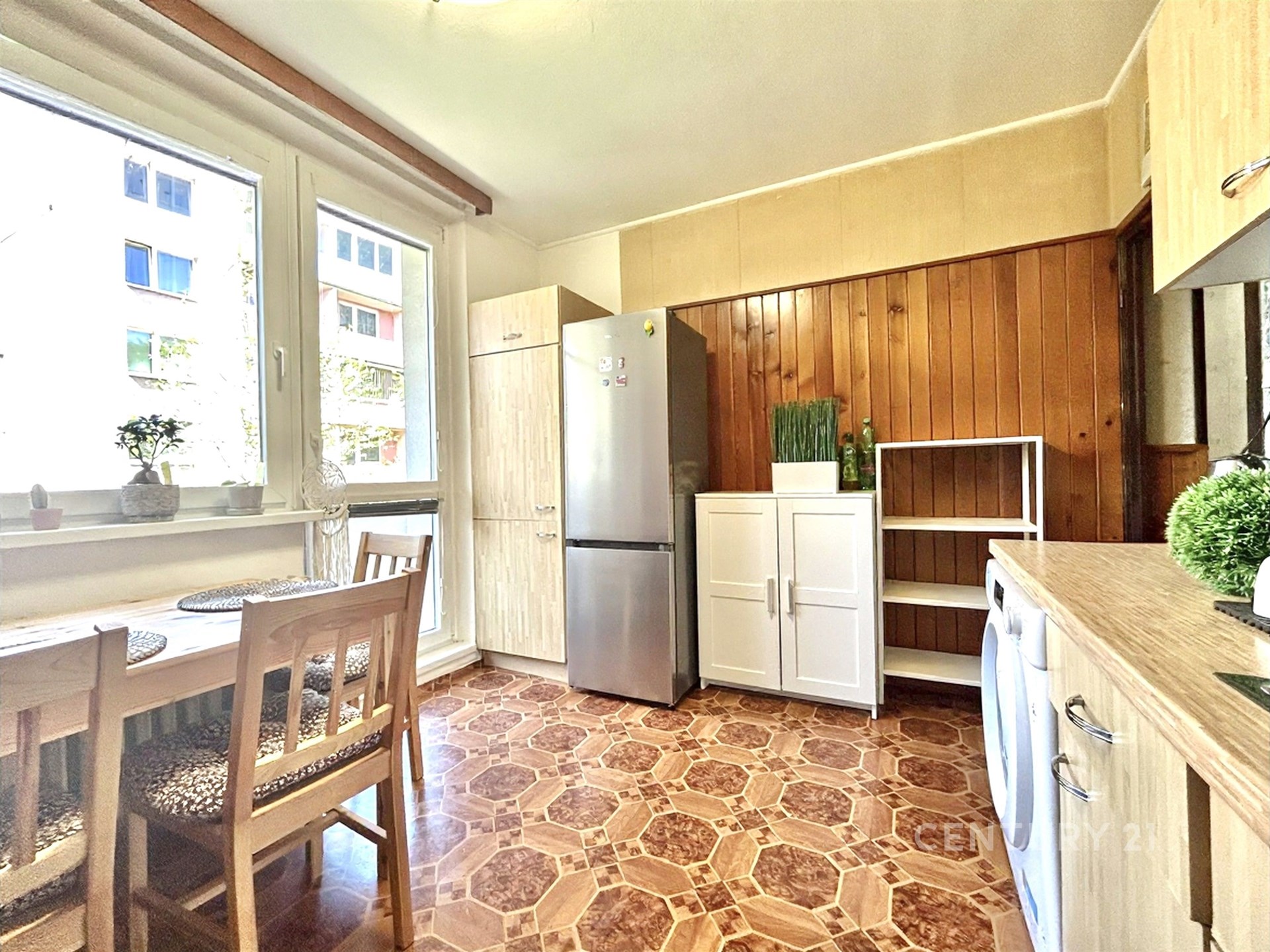 Světlá kuchyň s dřevěnými obklady, parketovou podlahou a velkými okny. Nábytek zahrnuje vestavěné skříně a moderní spotřebiče.