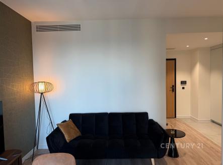 Světlý obývací pokoj s parketovou podlahou, moderní sedačkou a stylovým osvětlením, vedoucí do prostorné chodby s dlažbou. | Prodej bytu, 1+kk, 24 m²