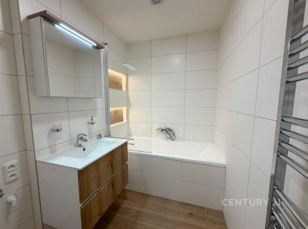 Moderní koupelna s bílou dlažbou, dřevěným umyvadlovým pultem a vanou. Světlý a čistý design s praktickým zrcadlem a topným žebříkem. | Pronájem bytu, 3+kk, 62 m²
