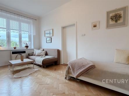 Světlý obývací pokoj s parketovou podlahou, velká okna zajišťují přirozené světlo, moderní dřevěný nábytek dodává elegantní vzhled. | Prodej bytu, 1+1, 43 m²