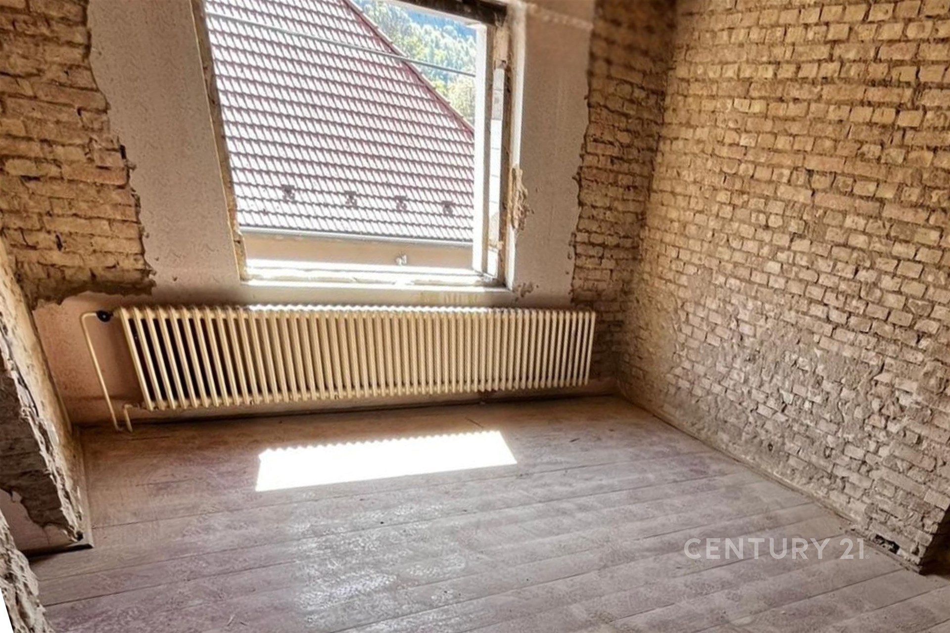 Pokoj ve fázi rekonstrukce s odkrytými cihlovými stěnami, světlým dřevěným podlahovým povrchem a velkým oknem pro přirozené světlo.