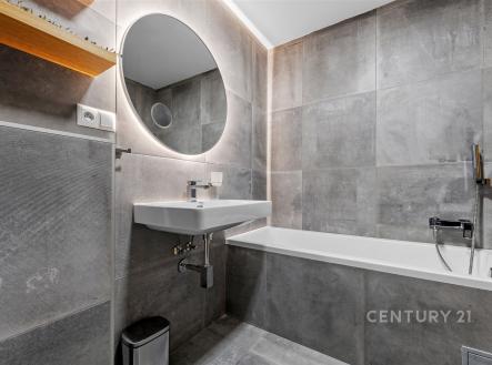 Moderní koupelna s šedými dlaždicemi, velkým kulatým zrcadlem a vestavěnou vanou. Stylové osvětlení a elegantní umyvadlo. | Prodej bytu, 3+kk, 68 m²
