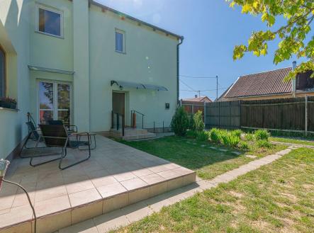 Zahrada s terasou se světlou dlažbou, vybavena venkovním posezením. Moderní oplocení nabízí soukromí, okolí doplňuje zeleň a květinové záhony. | Prodej - dům/vila, 210 m²