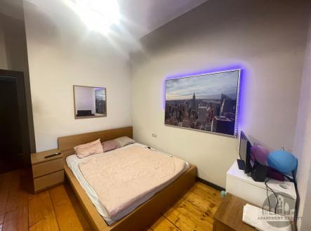 Prostorná ložnice s dřevěnou podlahou a moderním osvětlením, vybavená vestavěnou skříní a velkou postelí, nabízí komfort a styl. | Pronájem bytu, garsoniéra, 13 m²