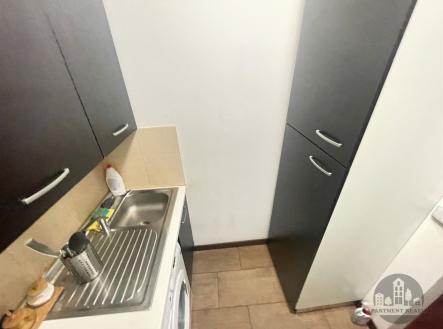 Kompaktní kuchyň s dlažbou a tmavými skříněmi, dobře osvětlená a s vestavěnými spotřebiči, působí moderně a funkčně. | Pronájem bytu, garsoniéra, 13 m²