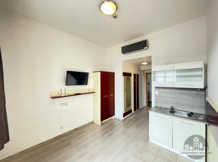 Kompaktní kuchyňský kout s moderními skříňkami, světlou dlažbou a parketovou podlahou propojený s obytným prostorem vybaveným vestavěnou skříní. | Pronájem bytu, 1+kk, 16 m²