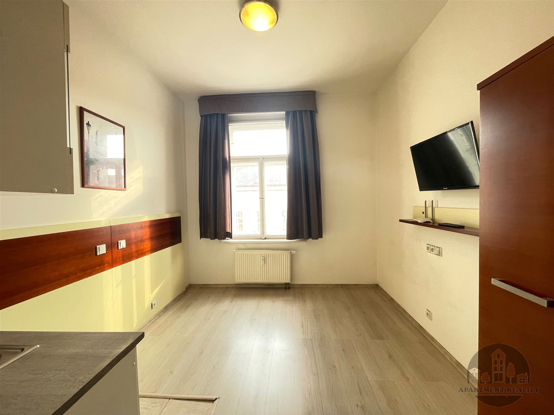 Světlý pokoj s moderní parketovou podlahou a velkým oknem, prostor je moderní a nabízí minimalistický design s nástěnnou televizí.