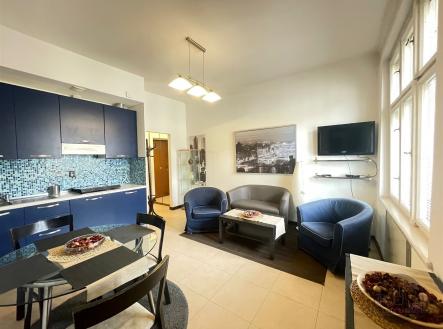 Kuchyňský kout s tmavě modrými skříňkami, světlá dlažba a moderní osvětlení, vedle obývací zóny s pohovkami a velkými okny. | Pronájem bytu, 2+kk, 37 m²