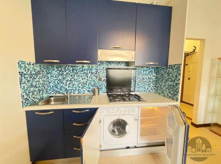Moderní kuchyň s modrými skříněmi, mozaikovým obkladem a vestavěnými spotřebiči, jako je pračka a plynový sporák, na světlé dlažbě. | Pronájem bytu, 2+kk, 37 m²