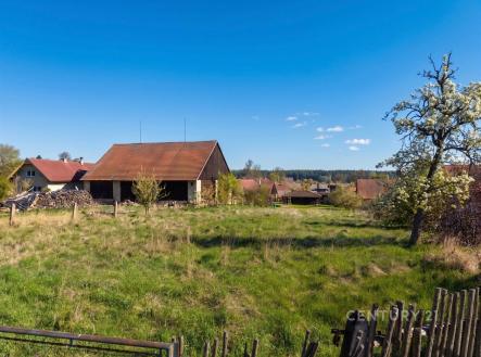 Prázdný stavební pozemek s travnatým porostem a dřevěným plotem, na okraji obce, s výhledem na okolní krajinu a stodolou v pozadí. | Prodej - pozemek pro bydlení, 604 m²