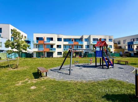 Dětské hřiště před moderním bytovým komplexem, obklopené zelení a lavičkami, nabízí klidné prostředí pro rodinné bydlení. | Prodej bytu, 3+kk, 67 m²