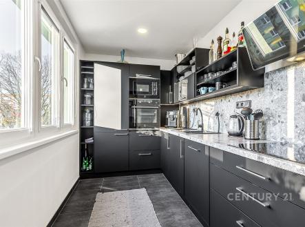 Moderní kuchyň s dlažbou, vestavěnými spotřebiči a velkými okny. Černé skříňky s kovovými úchyty dodávají elegantní vzhled. | Prodej bytu, 2+1, 57 m²