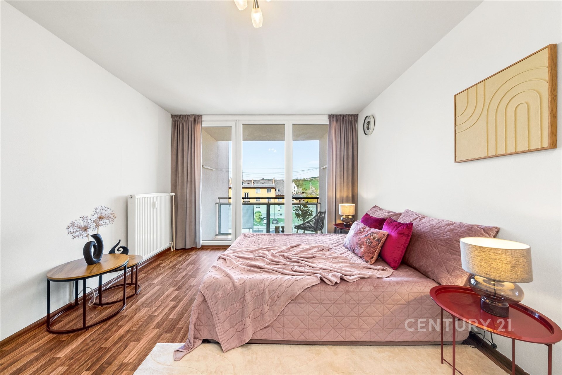 Světlá ložnice s parketovou podlahou, velká okna vedoucí na balkon, moderní dekorace a útulné uspořádání vytvářejí příjemné prostředí.