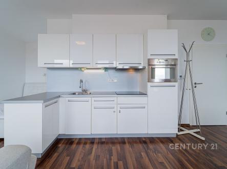 Moderní kuchyň s bílými lesklými skříňkami, vestavěnou troubou a parketovou podlahou, prostorný design pro pohodlné bydlení. | Pronájem bytu, 1+kk, 43 m²