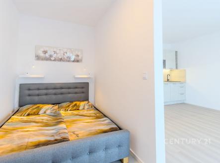Kompaktní ložnice s moderní šedou postelí, světlými stěnami a světlou laminátovou podlahou sousedící s otevřenou kuchyní. | Pronájem bytu, 1+kk, 43 m²