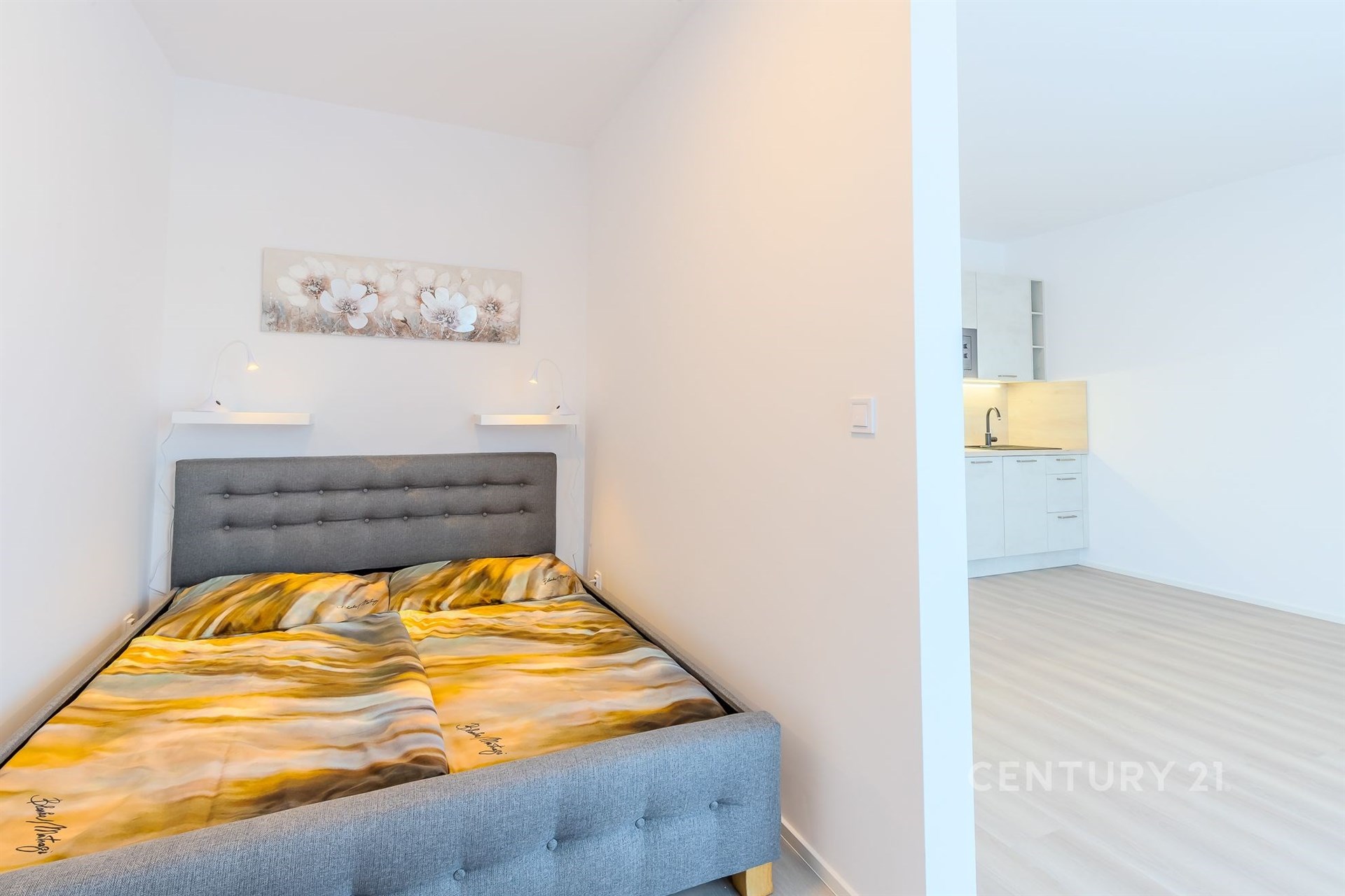 Kompaktní ložnice s moderní šedou postelí, světlými stěnami a světlou laminátovou podlahou sousedící s otevřenou kuchyní.
