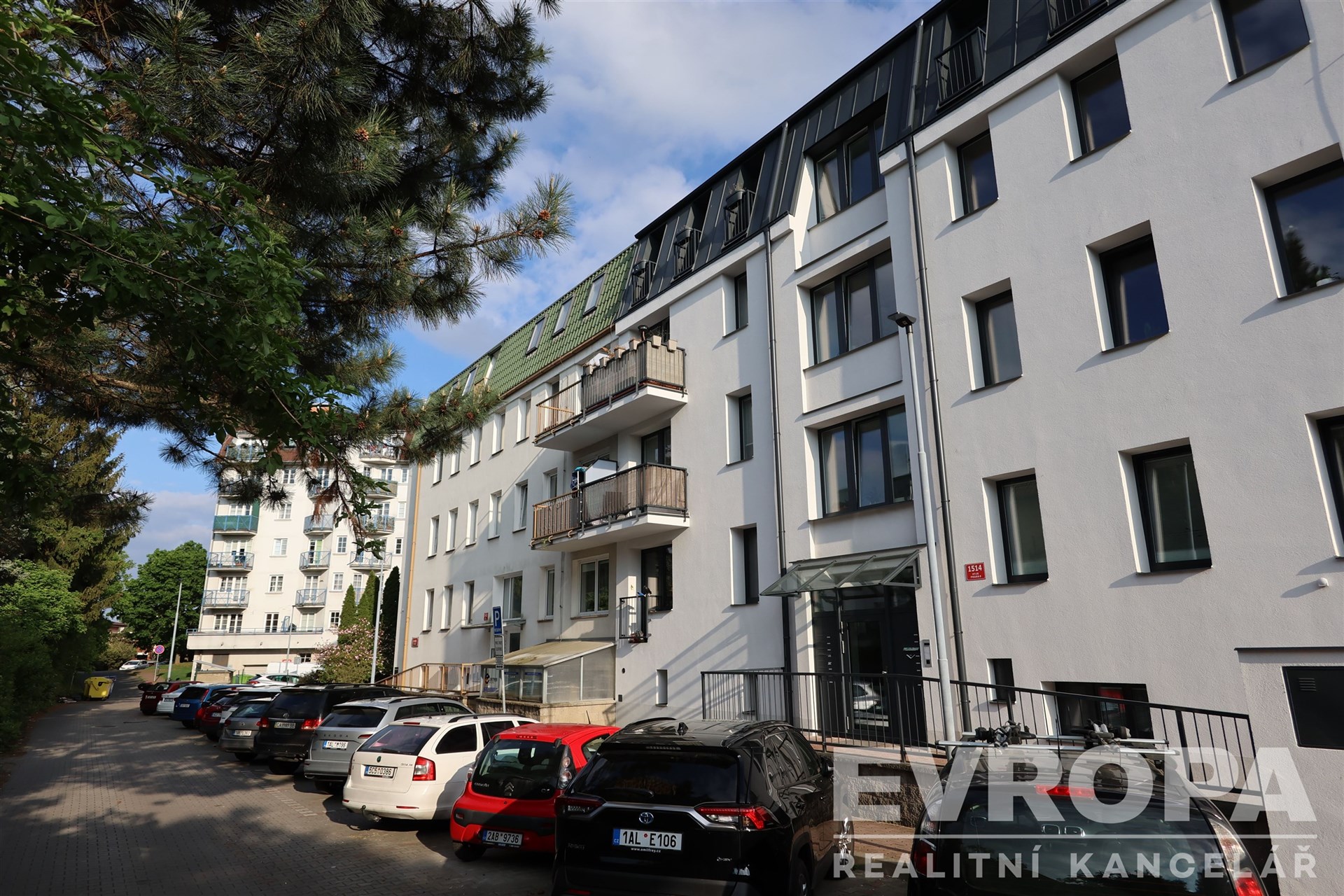 Moderní bytový dům, rekonstruovaný exteriér, bílá fasáda, velká okna, klidná rezidenční ulice, parkování před budovou.