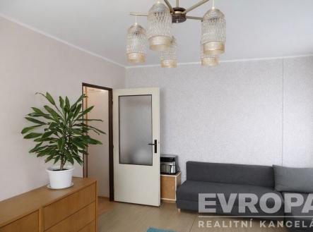 Světlý obývací pokoj s parketovou podlahou, moderní pohovka a designový lustr, interiér s neutrálními barvami a velkou rostlinou. | Prodej bytu, 2+1, 53 m²