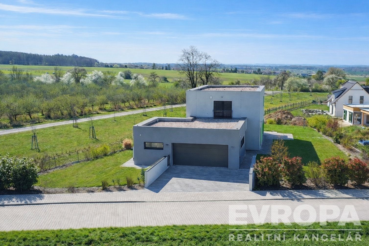 Moderní rodinný dům s plochou střechou a garáží, obklopený udržovanou zahradou, dlážděná příjezdová cesta, klidná lokalita blízko přírody.