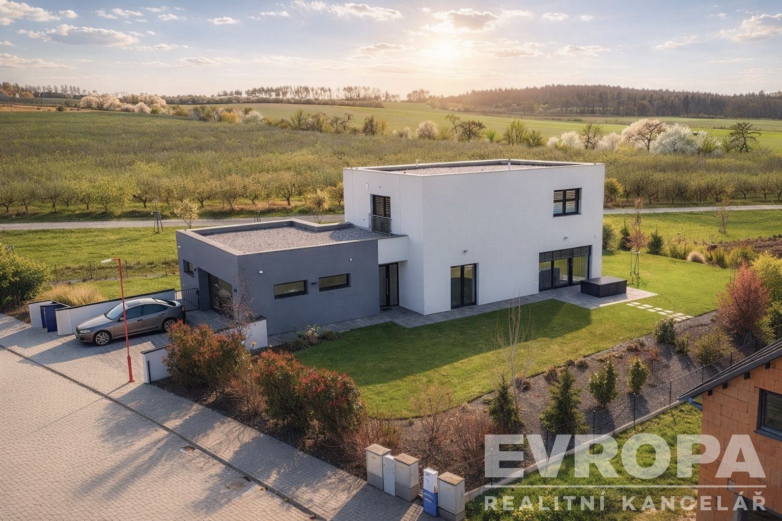 Moderní rodinný dům s plochou střechou, velká okna, zahrada a příjezdová cesta s dlažbou, nachází se v klidné přírodní lokalitě