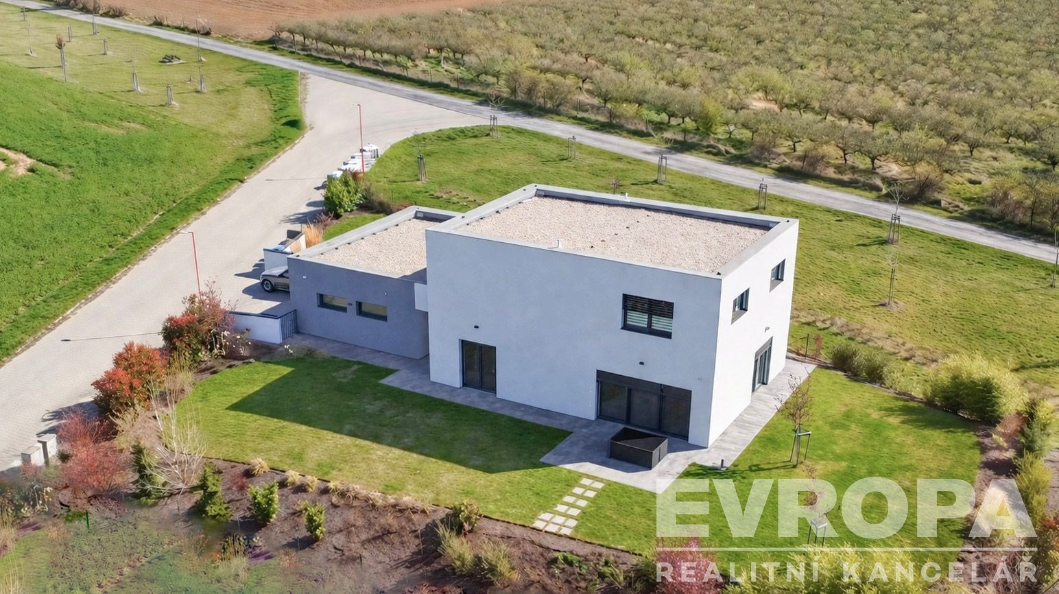 Moderní rodinný dům s plochou střechou, velkými okny a betonovou terasou, obklopený upravenou zahradou na klidném předměstí.