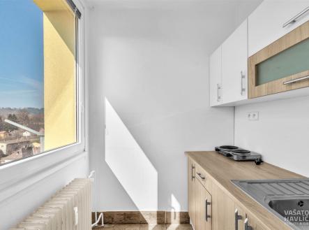 Malá kuchyň s moderními skříňkami, parketovou podlahou a velkým oknem poskytujícím přirozené světlo. | Pronájem bytu, 1+kk, 26 m²