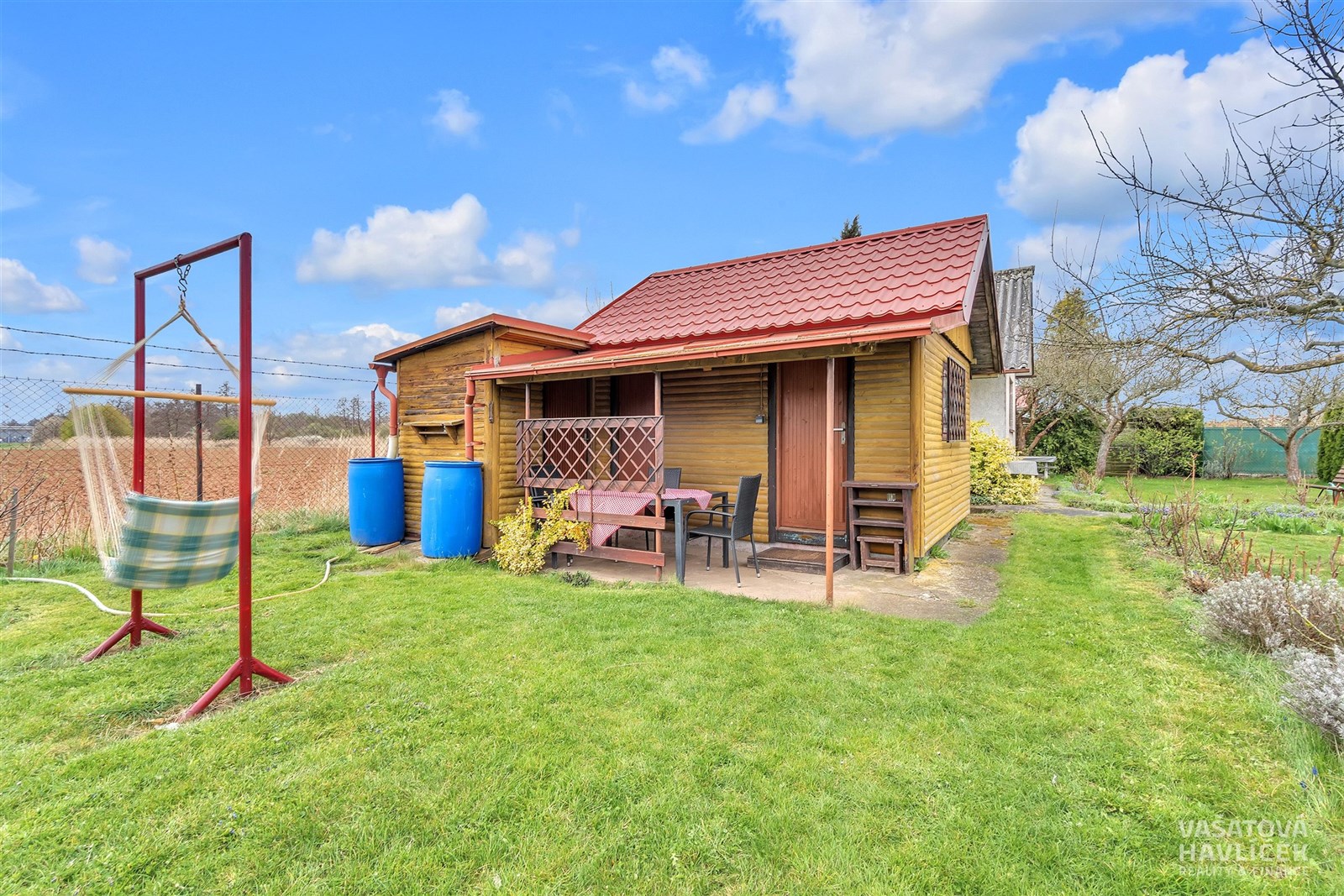 Malá chatka s dřevěnou fasádou, okolní zahrada s trávníkem a venkovním posezením, klidné prostředí na okraji vesnice.