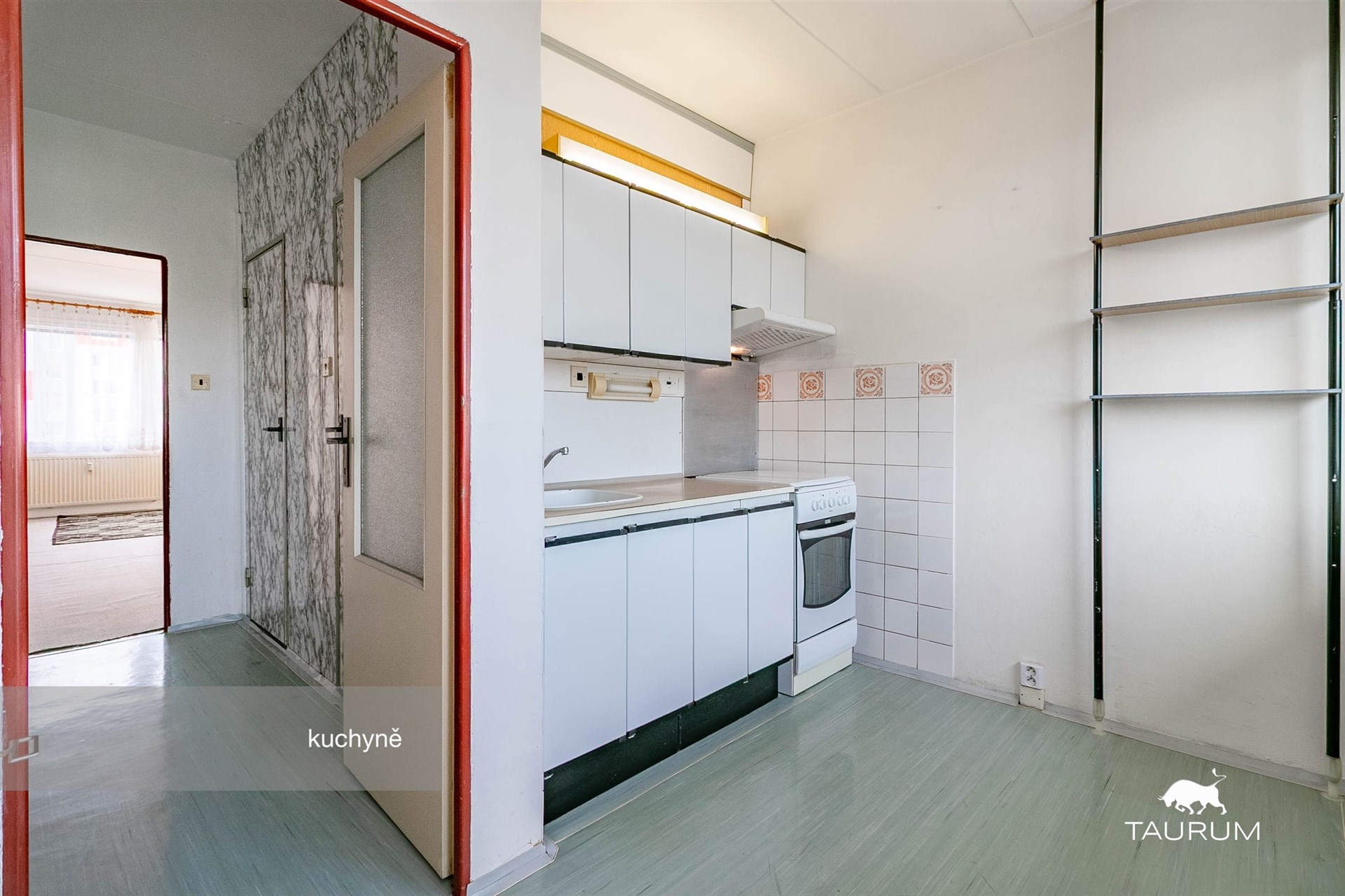 Kuchyň s bílými skříňkami a linoleem, světlý prostor s oknem. Obsahuje vestavěné police a dlažbu u sporáku, ideální pro modernizaci.