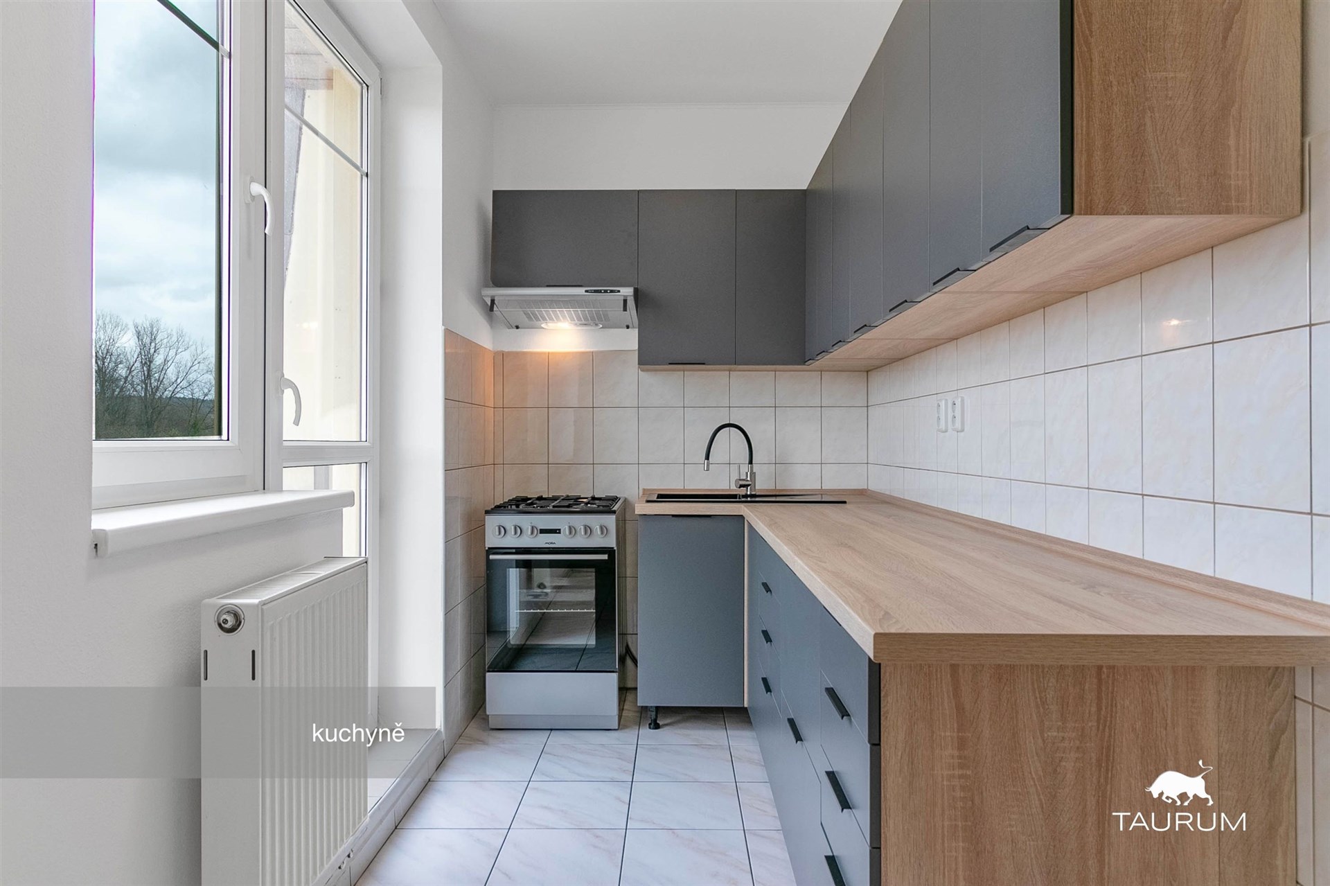 Moderní kuchyň s dlažbou na podlaze, dřevěnou pracovní deskou a vestavěnými skříněmi, velké okno zajišťuje dostatek světla.