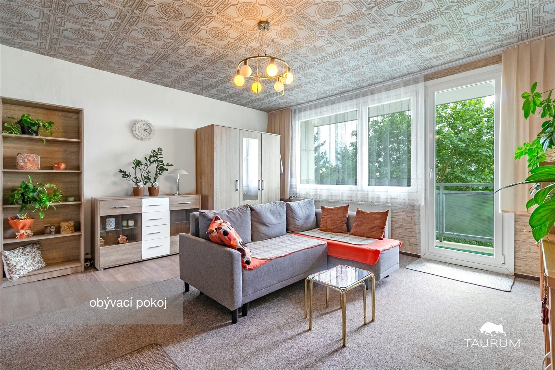 Světlý obývací pokoj, moderní styl, velká okna, dřevěný nábytek, renovované podlahy, přístup na balkon, svěží atmosféra.