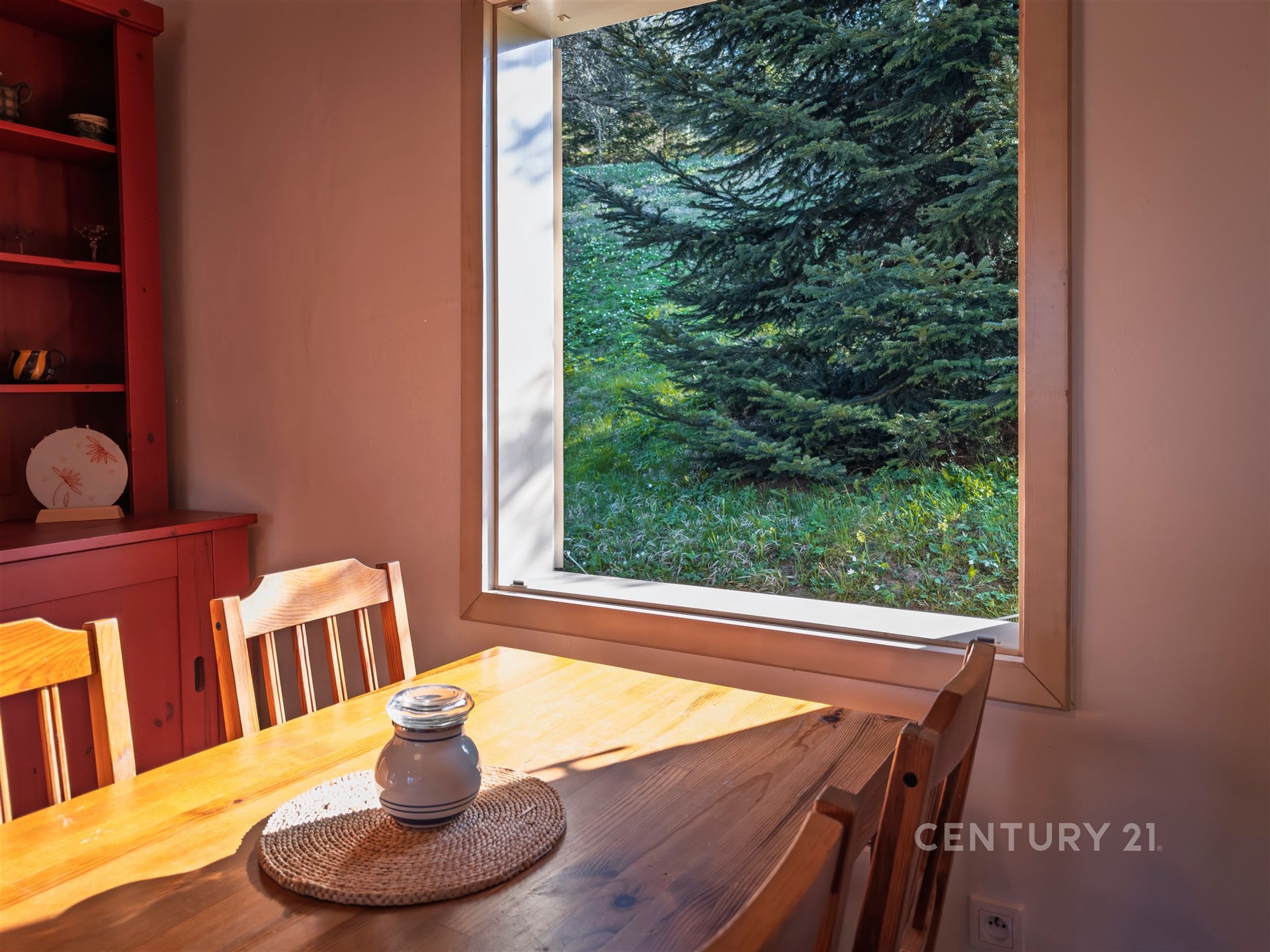 Jídelna s masivním dřevěným stolem, velké okno s výhledem na zahradu, přírodní světlo a útulný interiér.