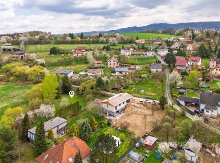 Letecký pohled na vesnickou oblast s rozptýlenými domy a zahradami obklopenými zelení, s výhledem na kopcovitou krajinu v pozadí. | Prodej - pozemek pro bydlení, 1 904 m²