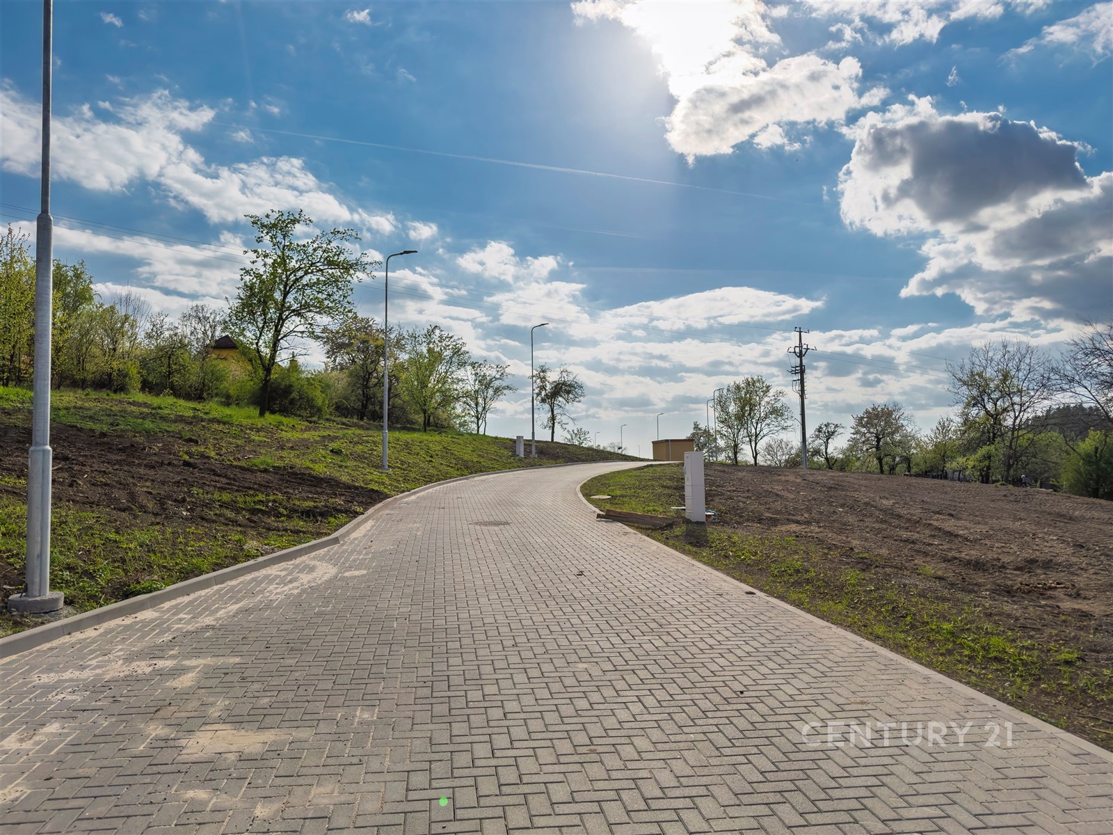 Nová zpevněná příjezdová cesta s dlážděným povrchem, obklopená zelení a stromovím, vytváří moderní a klidné okolí pro budoucí stavební parcelu.