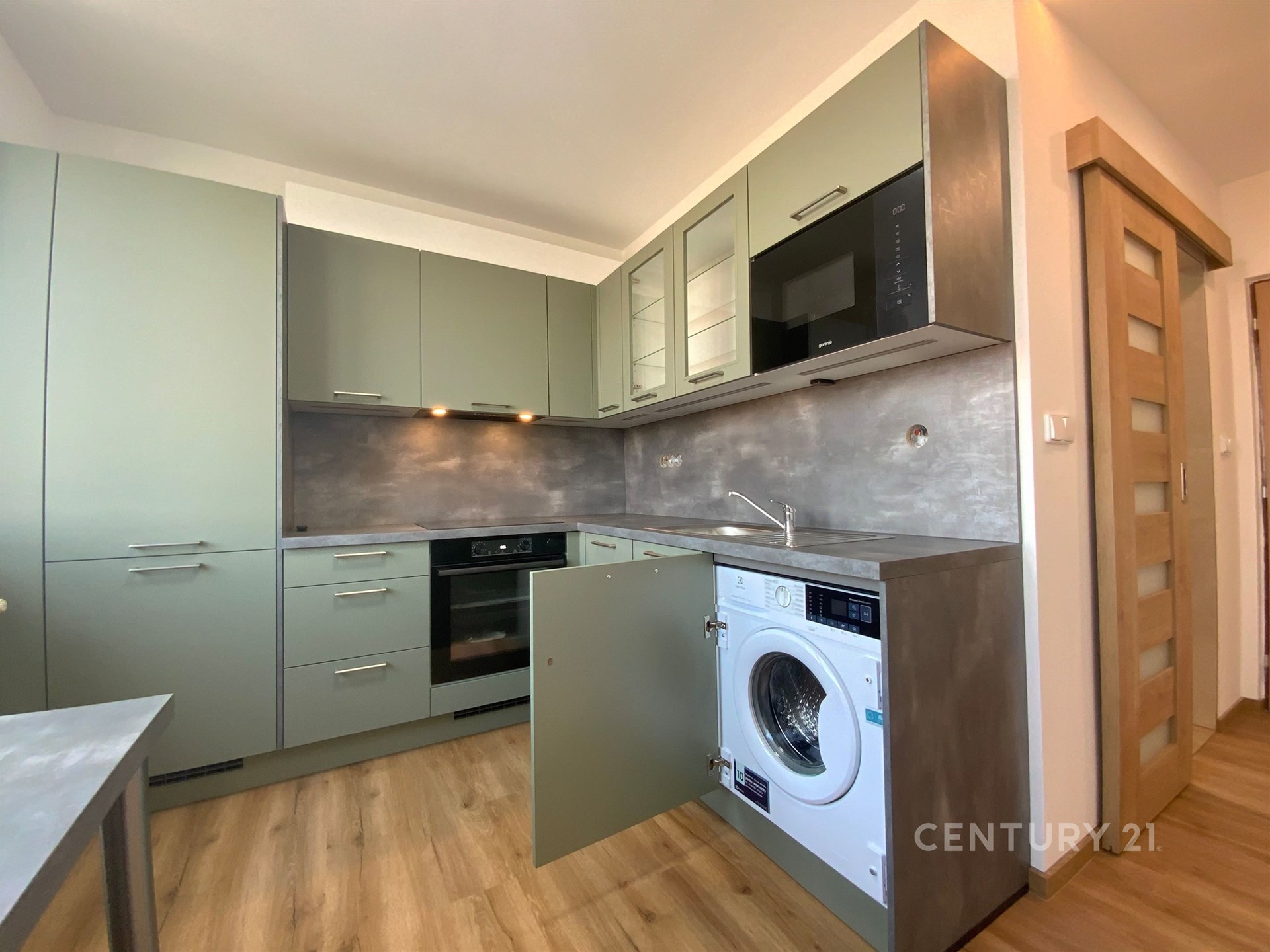 Moderní kuchyň s vestavěnými spotřebiči, parketovou podlahou a elegantními skříňkami v zelenošedém odstínu.