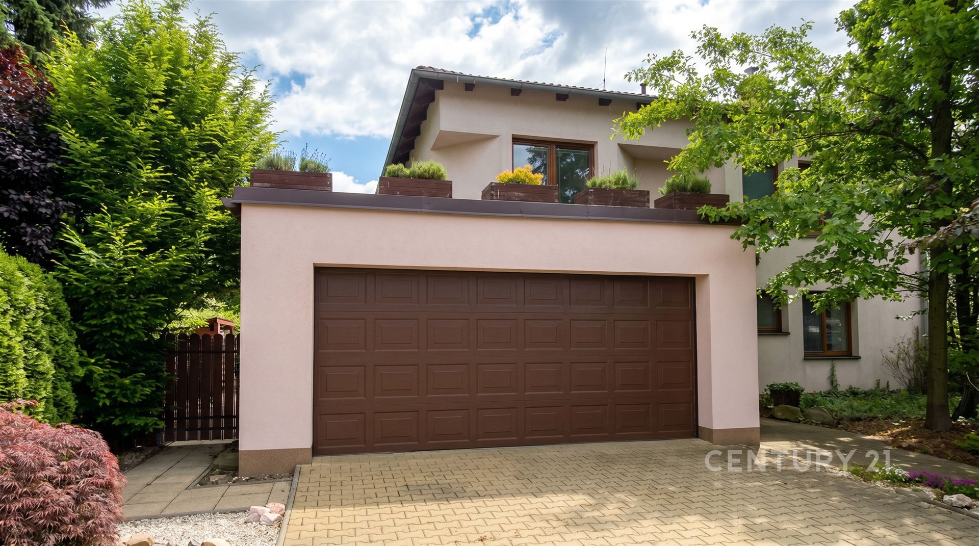 Dvojgaráž s hnědými vraty a zámkovou dlažbou, obklopena zelení. Moderní fasáda domu s terasou, která dodává příjemný dojem.