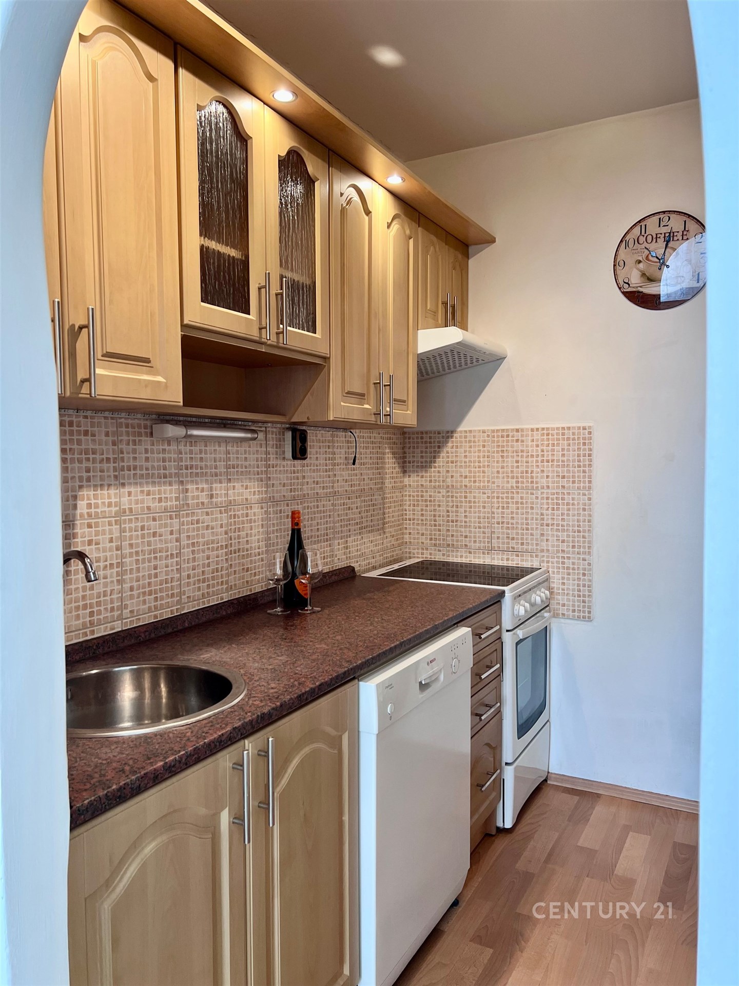 Kompaktní kuchyň s dřevěnými skříňkami, moderními spotřebiči a parketovou podlahou poskytuje praktický prostor pro vaření.