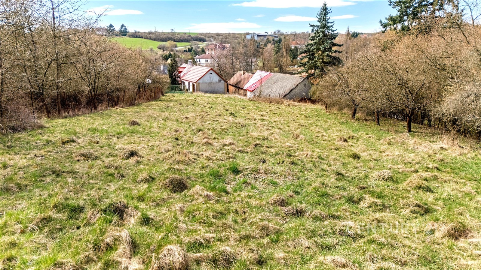 Stavební pozemek s travnatým porostem, obklopený přírodou a stromy, s výhledem na okolní venkovské domy, ideální pro rekreační stavbu.