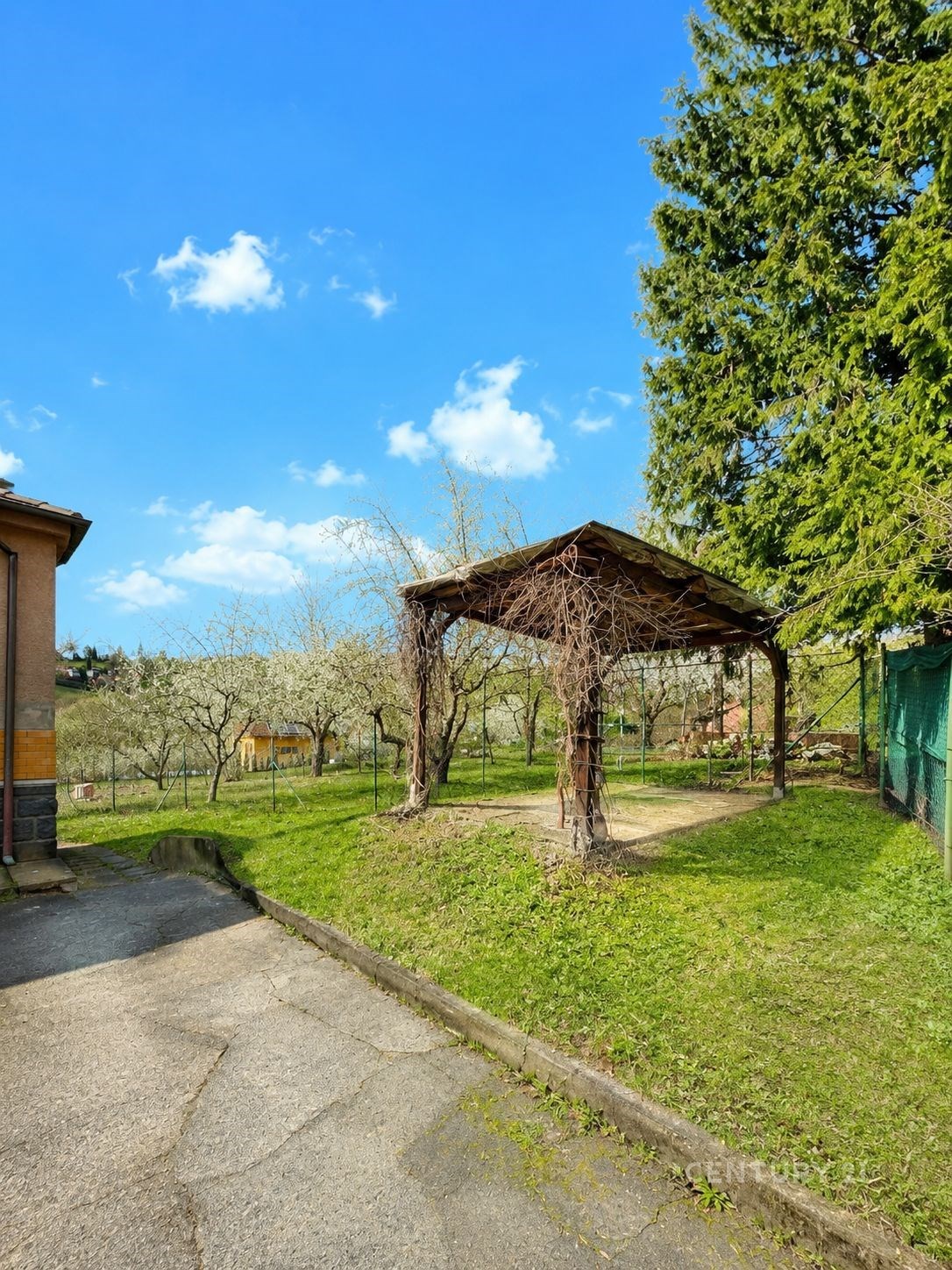 Zahrada s pergolou, částečně zastíněná dřevěnou konstrukcí. Přilehlá upravená travnatá plocha se vzrostlými stromy poskytuje příjemné prostředí.