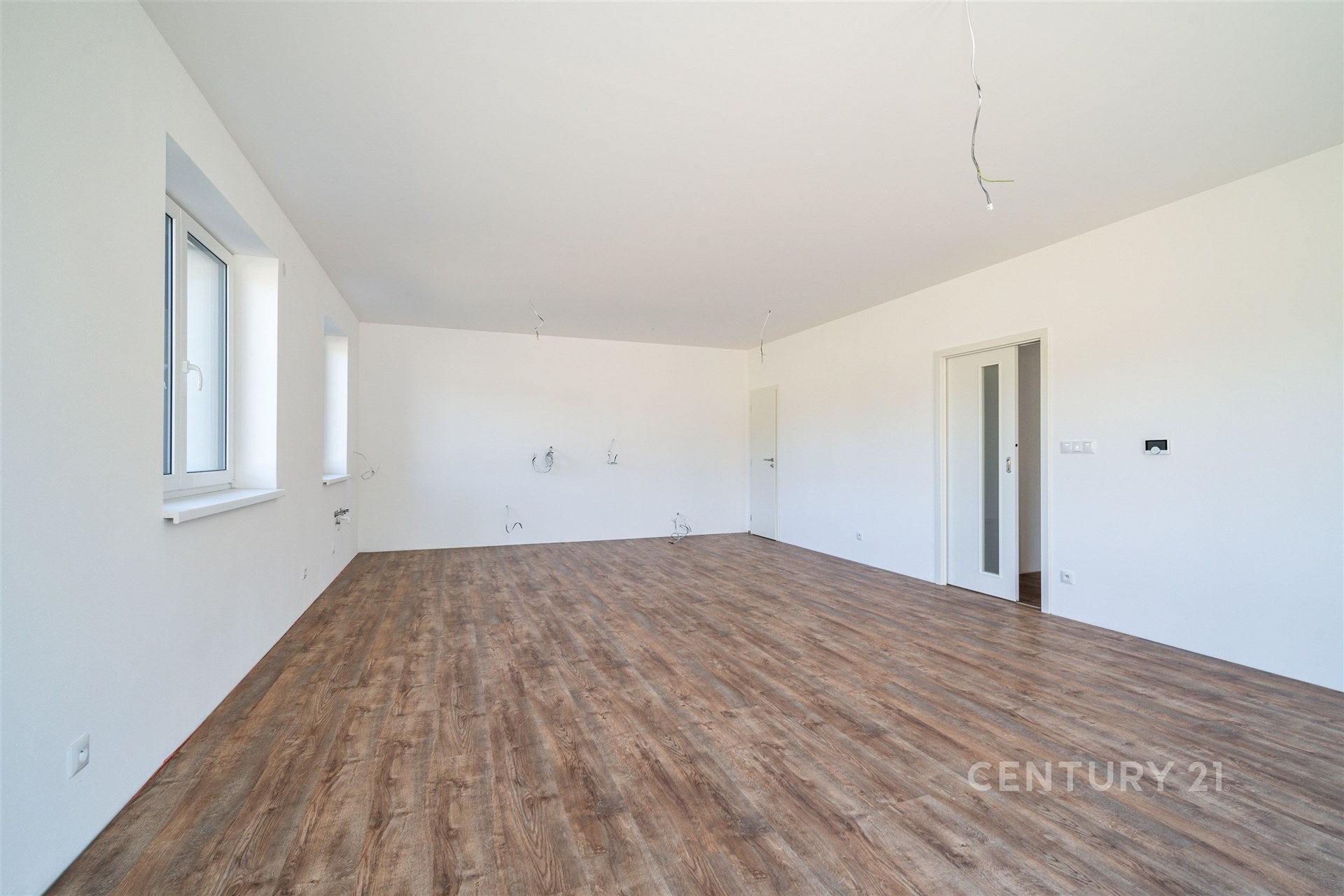 Prostorný obývací pokoj s parketovou podlahou, velká okna zajišťují dostatek světla, moderní minimalistický design v neutrálních barvách.