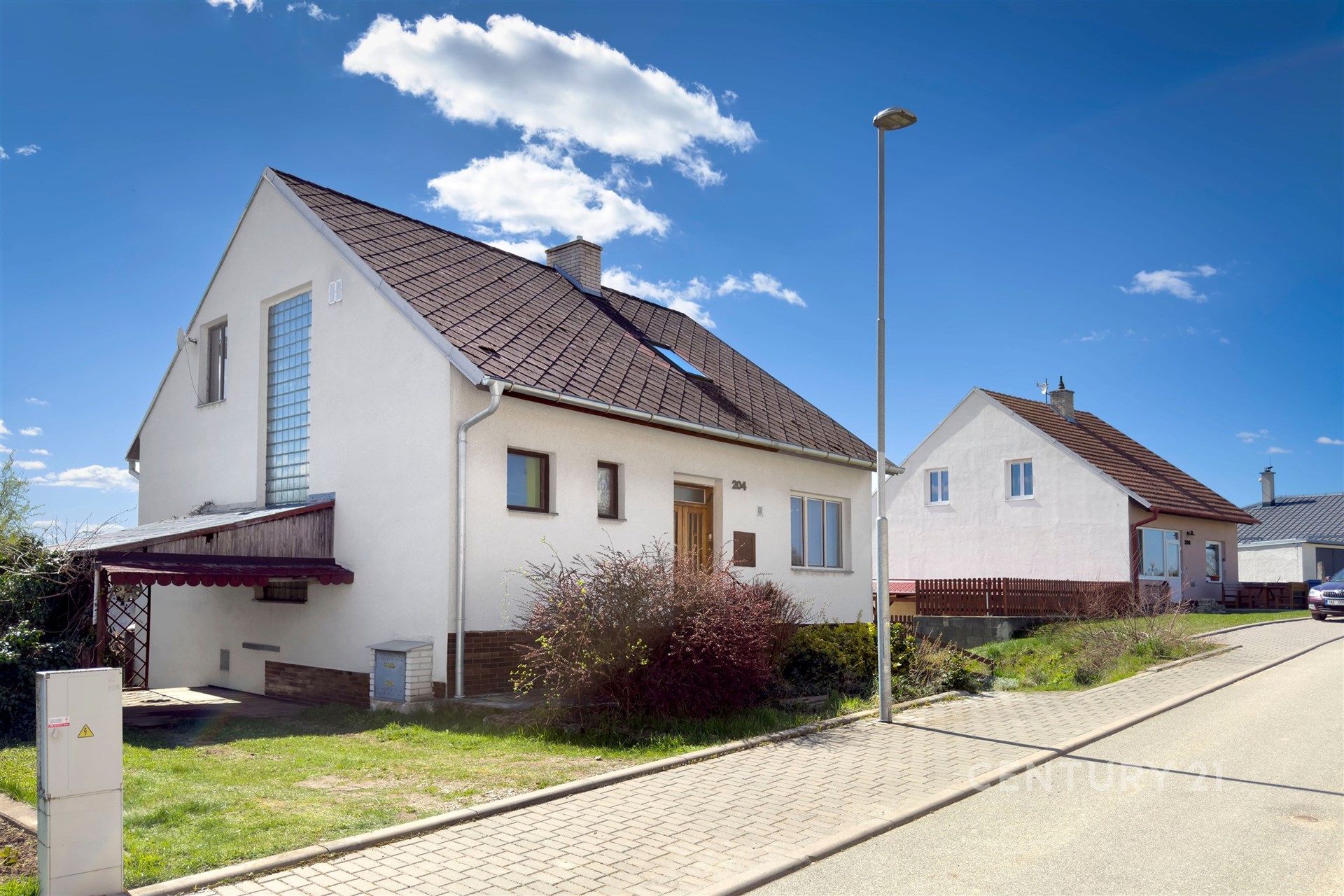 Rodinný dům s moderní fasádou, šikmou střechou a velkými okny, nacházející se na klidné ulici s dlážděným chodníkem.