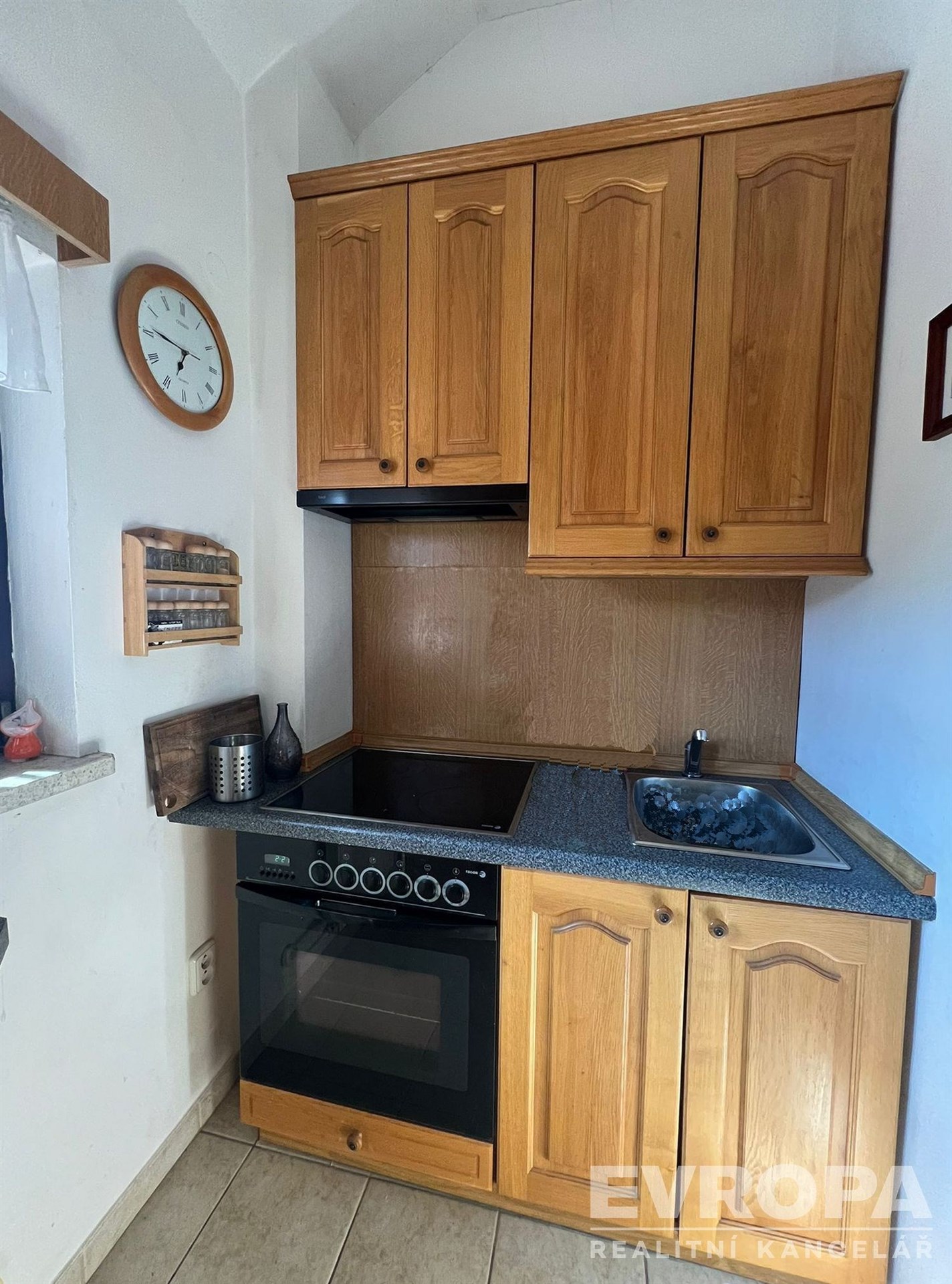 Malá kuchyň s dřevěnými skříněmi, elektrickým sporákem a dlažbou. Má praktické uspořádání a je ideální pro menší prostory.