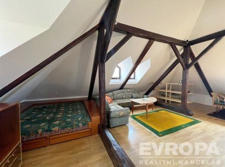 Prostorná podkrovní místnost s parketovou podlahou, dřevěnými trámy a velkými okny, vytvářející světlý a útulný prostor | Pronájem bytu, garsoniéra, 30 m²