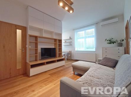 Světlý obývací pokoj s moderní dřevěnou podlahou, vestavěnou knihovnou a velkými okny, poskytující útulný a komfortní prostor. | Pronájem bytu, 2+kk, 54 m²