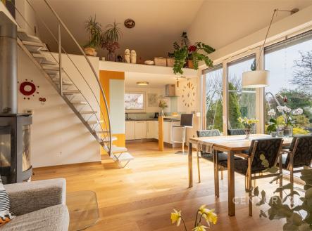 Světlý obývací pokoj s dřevěnou podlahou a velkými okny propojený s moderní kuchyní, krbová kamna vytvářejí útulnou atmosféru. | Prodej - dům/vila, 115 m²