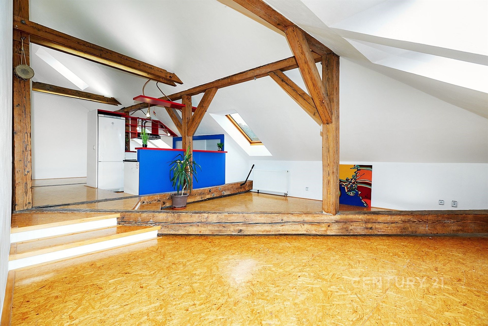 Podkrovní prostor s dřevěnými trámy, parketovou podlahou a moderními prvky, nabízí světlý interiér s vestavěnými doplňky a uměleckým akcentem.