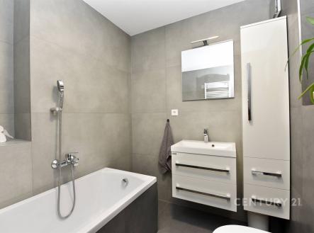 Moderní koupelna s dlažbou, bílými skříňkami a zrcadlem nad umyvadlem, nabízí elegantní a funkční interiér pro pohodlné bydlení. | Prodej - dům/vila, 273 m²