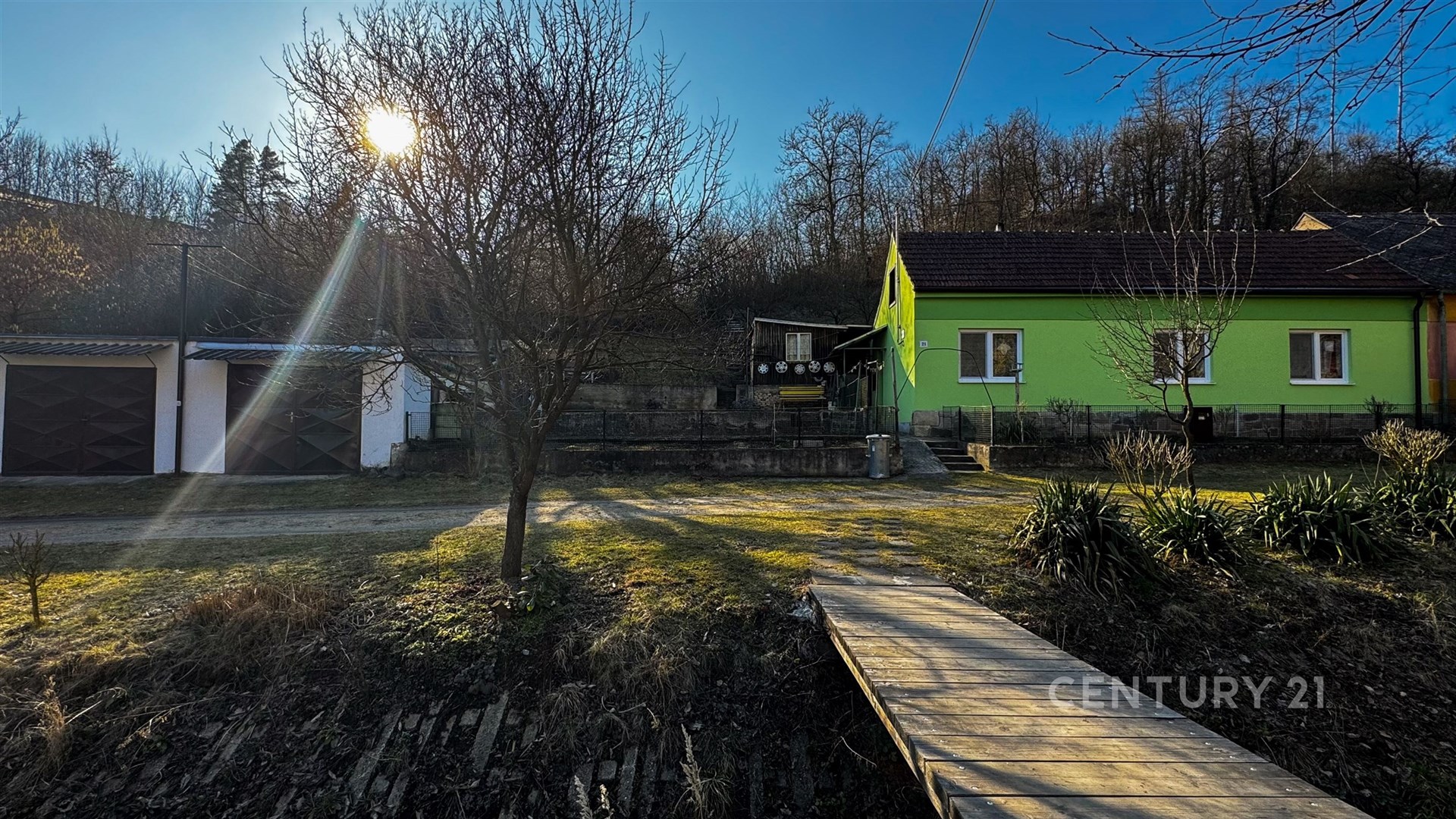 Zahrada s dřevěným chodníkem před zeleným rodinným domem, dvě garáže, klidná lokalita, ideální pro rodiny hledající venkovské bydlení.