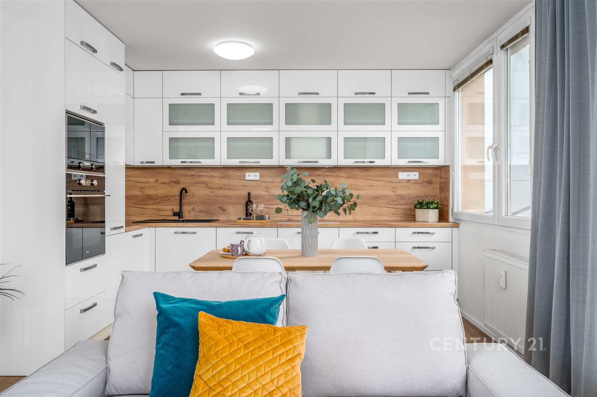 Moderní kuchyň s bílou vestavěnou linkou, dřevěnou pracovní deskou a parketovou podlahou, velká okna propouštějí dostatek světla.