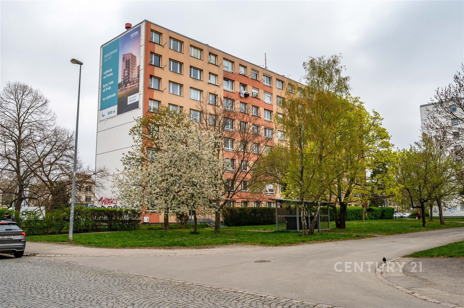 Vícepatrový bytový dům s moderní fasádou, obklopený zelení, stromy a parkovými plochami, vytváří příjemné městské prostředí.